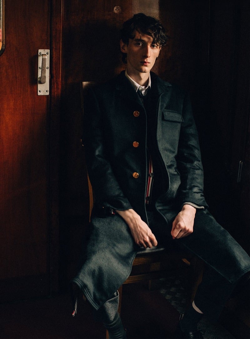 Arran Young sits in a mahogany-paneled train compartment wearing a long dark Thom Browne brass-button overcoat over a cardigan and white shirt, a red, white, and blue grosgrain tab at the chest