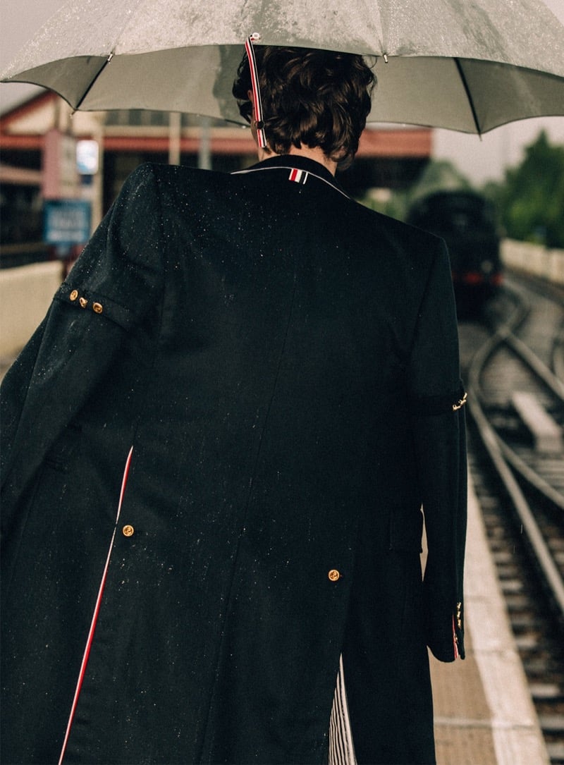 Arran Young seen from behind under a clear dome umbrella on a wet railway platform, wearing a long dark Thom Browne overcoat with gold cuff buttons and red, white, and blue grosgrain trim, a steam train approaching