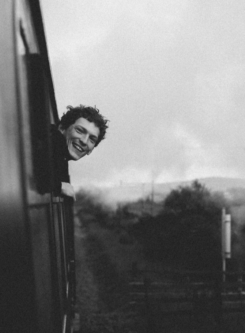 Black-and-white photo of Arthur del Beato leaning out of a moving train window, smiling broadly, wearing a dark Thom Browne coat, hills and steam behind him