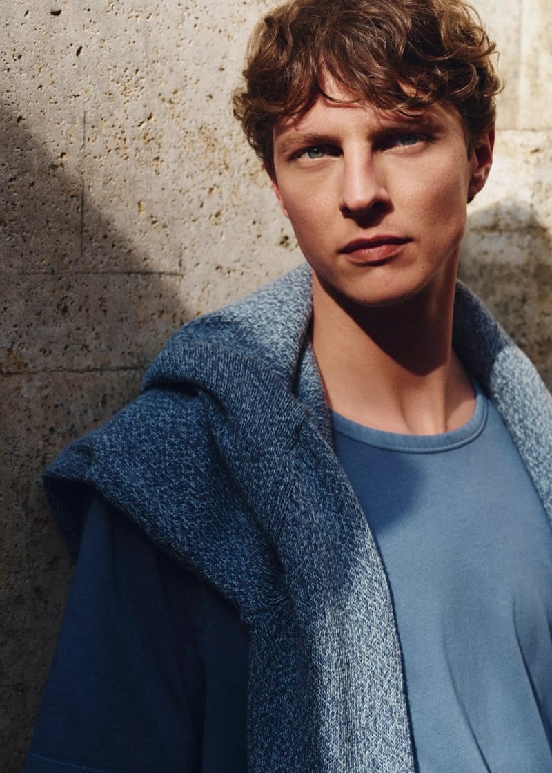 Tim Schuhmacher leans against a stone wall with a cable-knit sweater draped over his shoulders atop a blue T-shirt, for Mango's Superior Denim collection