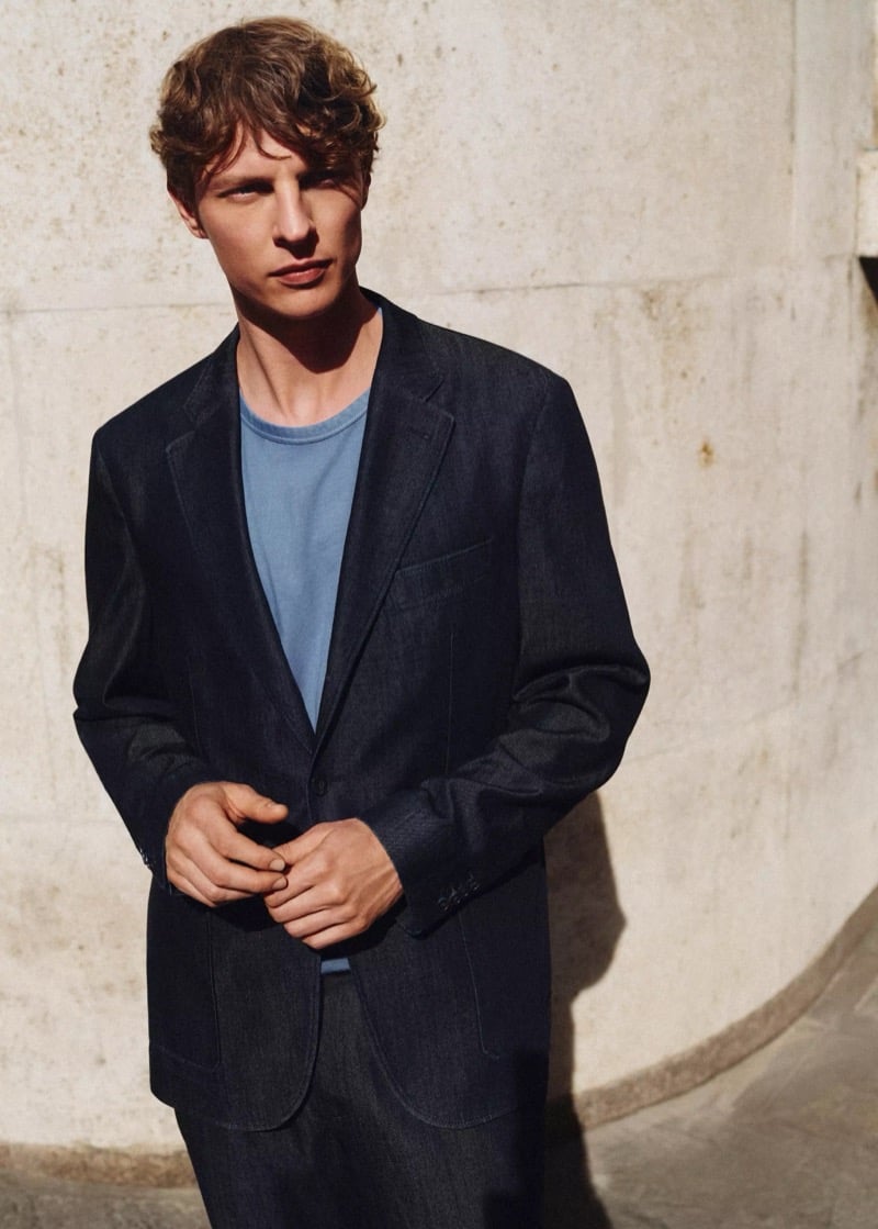 Tim Schuhmacher stands against a stone wall in an indigo denim blazer over a pale blue T-shirt with dark trousers, hands clasped, for Mango's Superior Denim collection