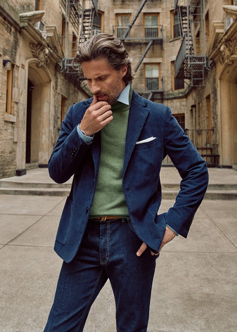 Tommy Dunn with a hand at his chin in a blue blazer, green turtleneck, and dark jeans in a brick courtyard with fire escapes