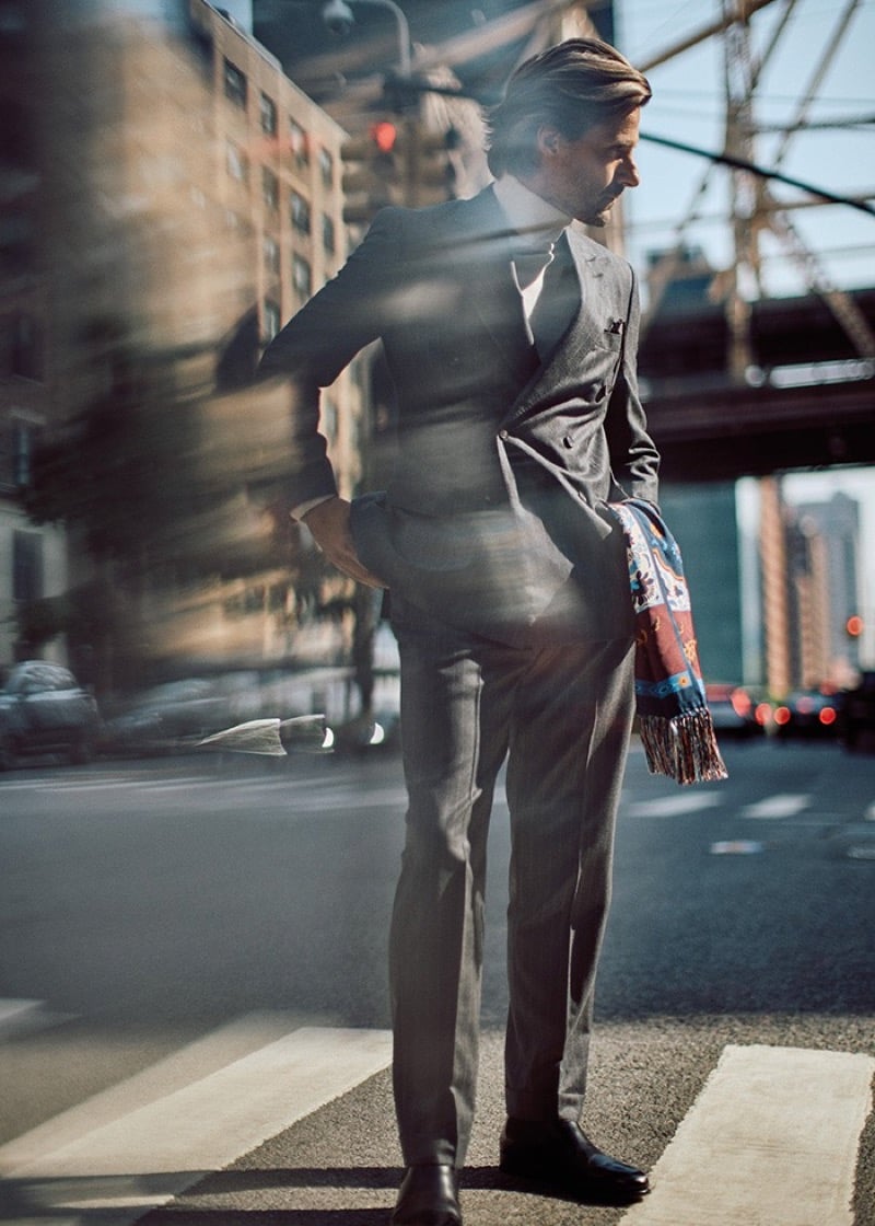 Tommy Dunn standing on a crosswalk in a gray double-breasted suit with a colorful fringed scarf in hand and city traffic blurred behind him