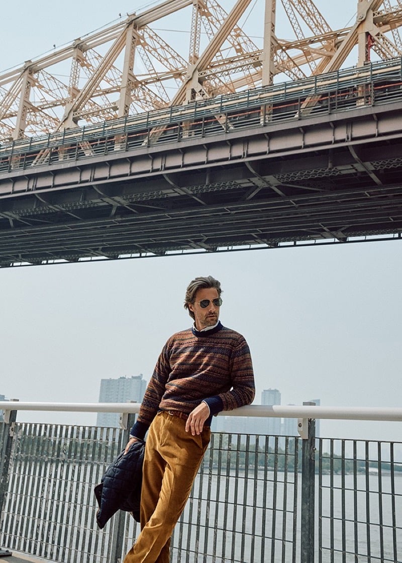 Tommy Dunn leaning on a waterfront railing under a bridge in aviator sunglasses, a striped patterned sweater, and mustard corduroys