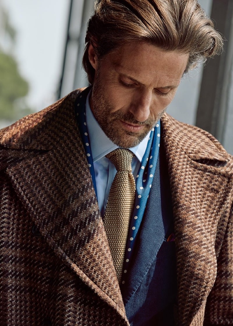 Tommy Dunn looking down in a brown plaid coat over a corduroy jacket with a light blue shirt, gold knit tie, and polka dot scarf