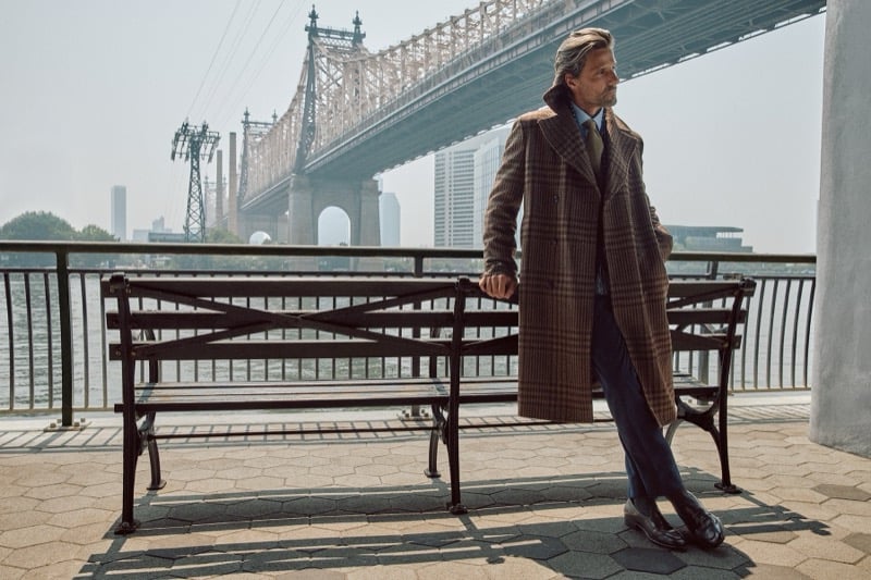Tommy Dunn leaning against a park bench in a long brown plaid overcoat and leather shoes with a bridge behind him