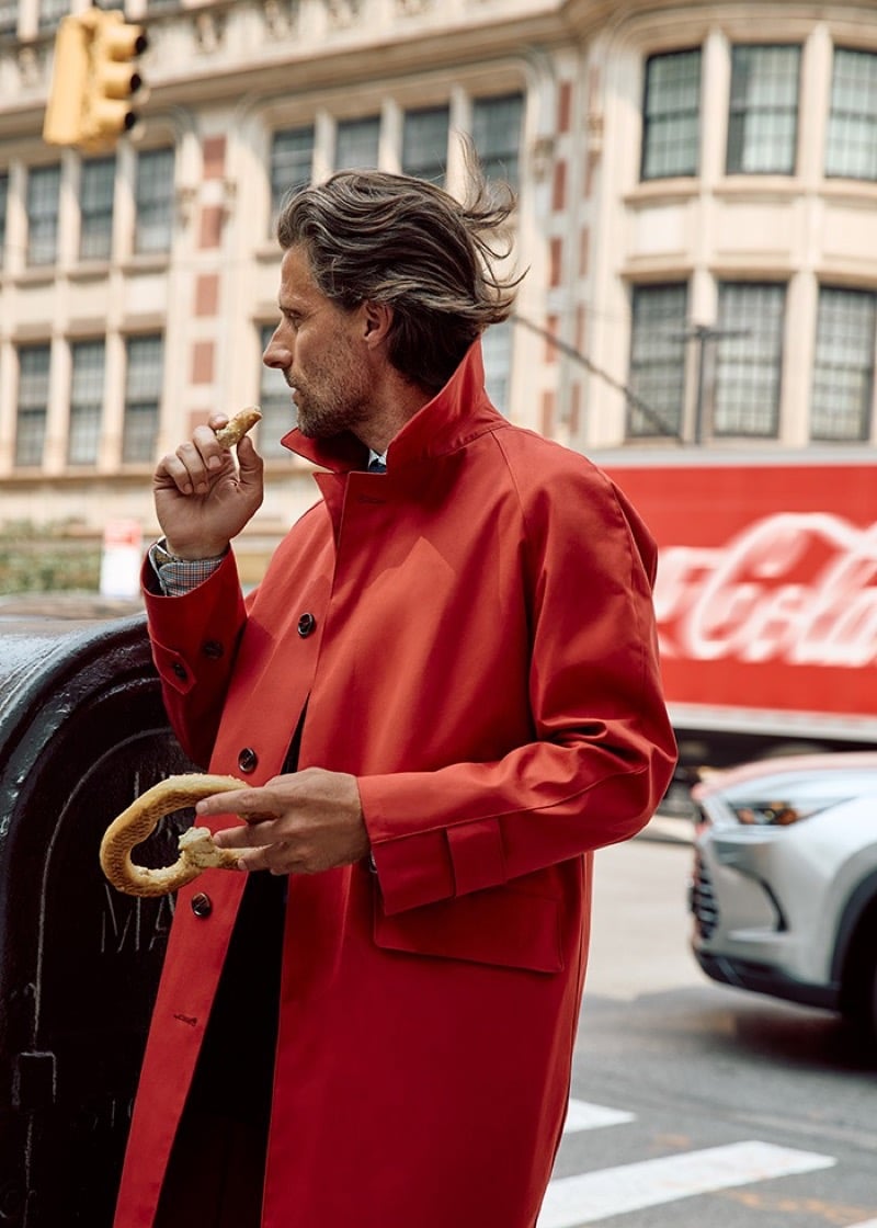 Tommy Dunn in a red button-front coat eating a pretzel beside a mailbox on a city street with a delivery truck behind him
