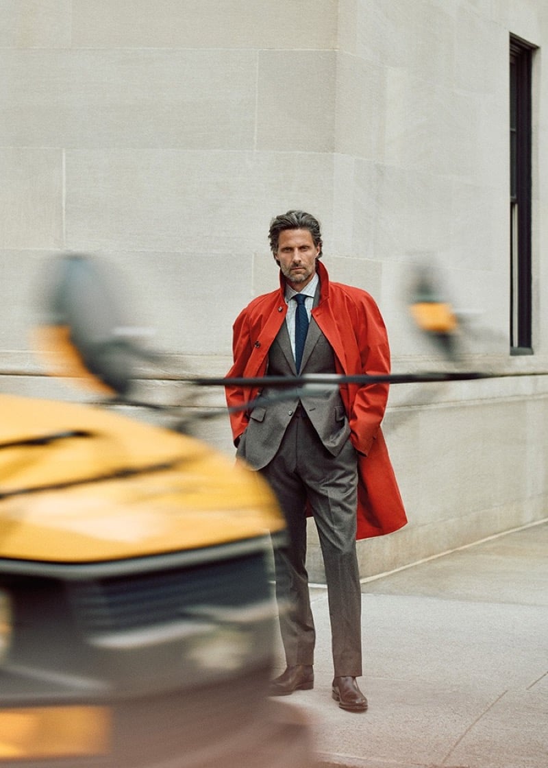 Tommy Dunn standing with his hands in the pockets of a red coat worn over a gray suit as a blurred taxi passes in the foreground