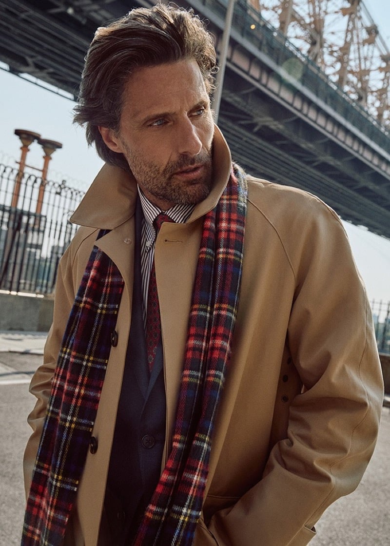 Close-up of Tommy Dunn in a tan raincoat with a red tartan scarf, striped shirt, and patterned tie beneath a bridge
