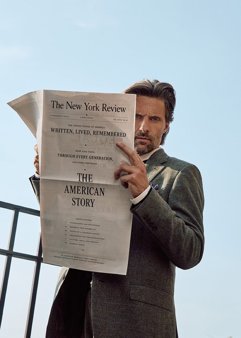 Tommy Dunn peering over a folded newspaper against a clear sky in a textured green-gray sport coat