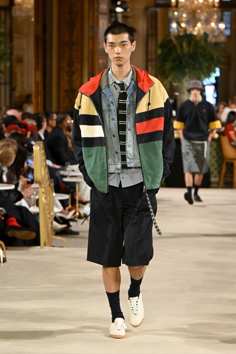 A model in a colorblock hooded jacket layered over a denim jacket and striped tie, with shorts and white sneakers, for Tommy Hilfiger's spring 2027 collection