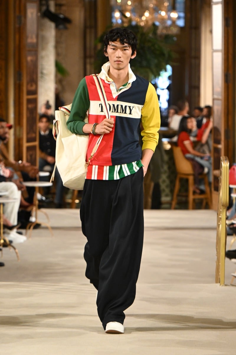 A model in a color-blocked rugby polo with a 'Tommy' wordmark, wide-leg trousers, and a canvas tote bag, for Tommy Hilfiger's spring 2027 collection