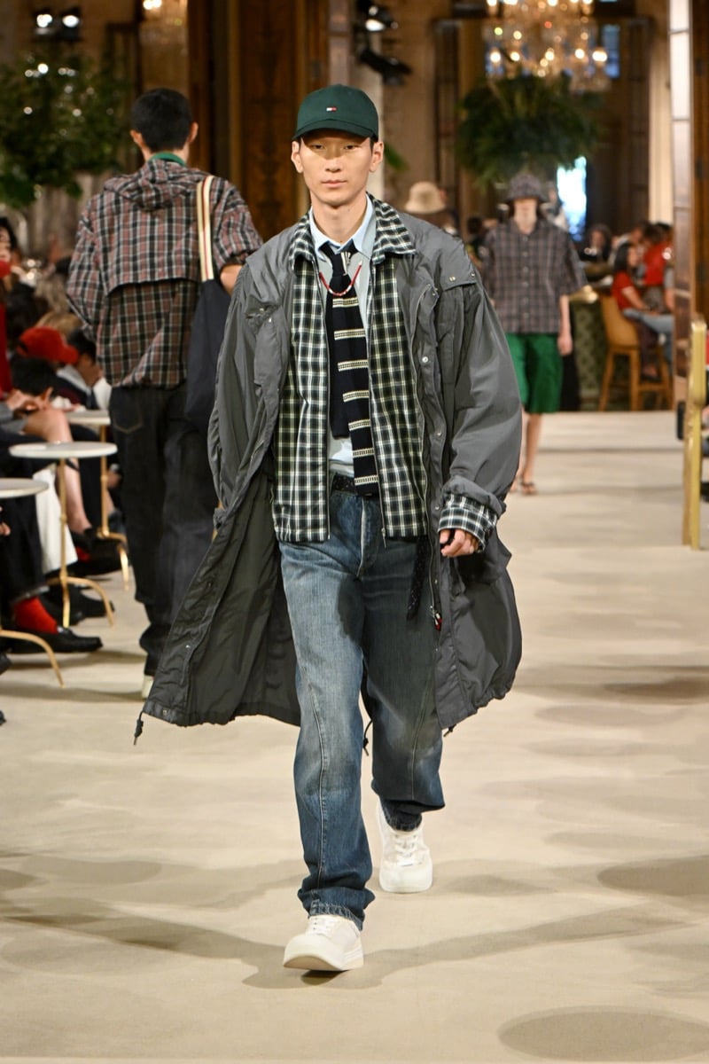 A model in a green cap and gray parka over a plaid shirt with a striped tie, paired with dark denim jeans, for Tommy Hilfiger's spring 2027 collection