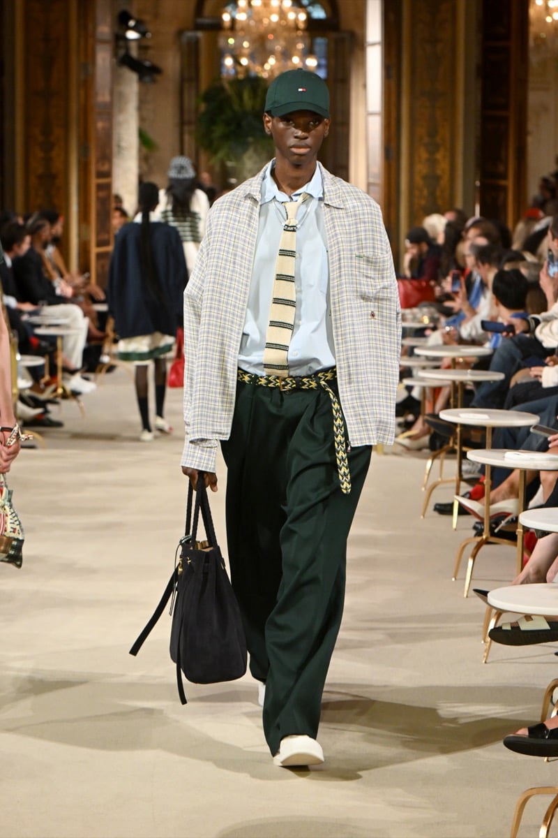 A model in a green cap and plaid overshirt with a striped tie, dark green trousers, and a bucket bag, for Tommy Hilfiger's spring 2027 collection