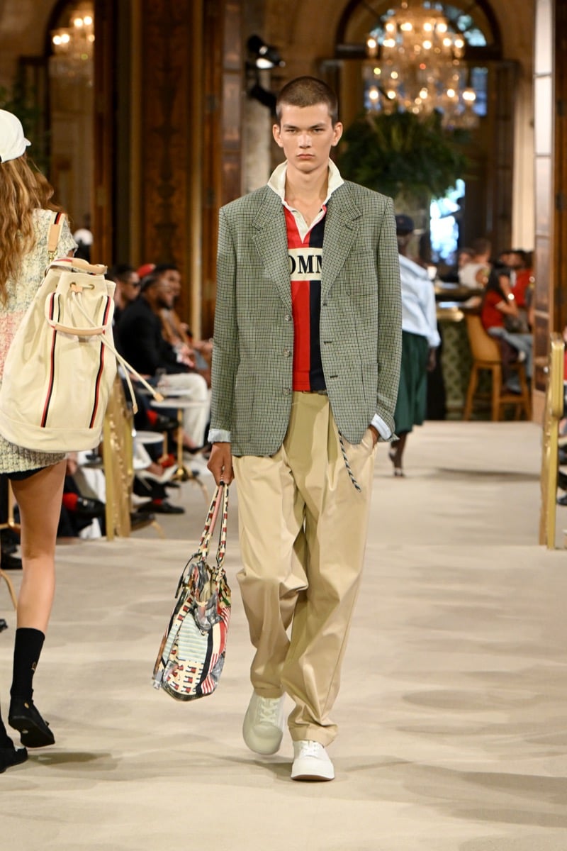 A model in a plaid blazer over a colorblock wordmark polo, carrying a flag-print tote bag with khaki trousers, for Tommy Hilfiger's spring 2027 collection
