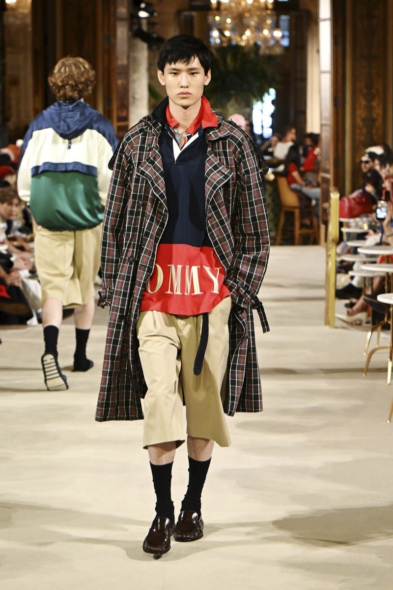 A model in a plaid trench coat over a colorblock wordmark polo, with khaki shorts and brown loafers, for Tommy Hilfiger's spring 2027 collection