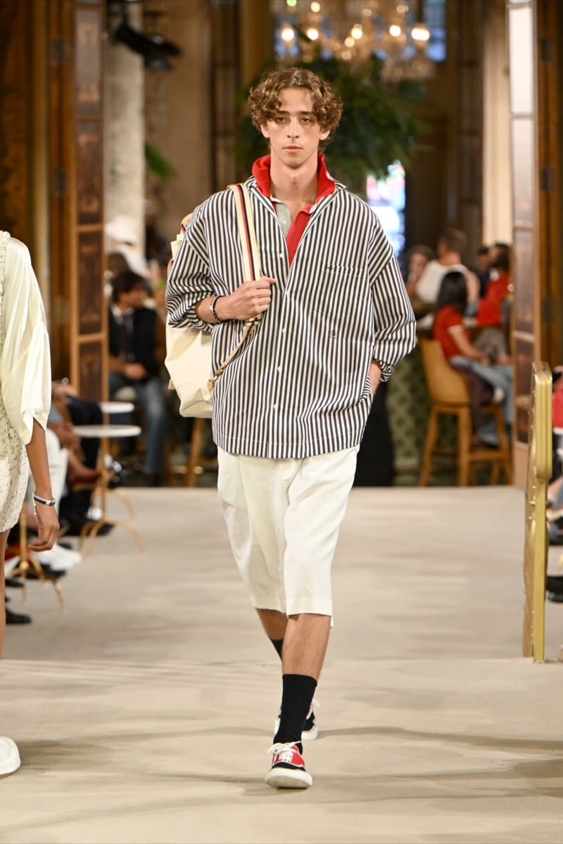 A model in an oversized striped shirt over a red collar, with a striped-strap tote bag and light-colored shorts, for Tommy Hilfiger's spring 2027 collection