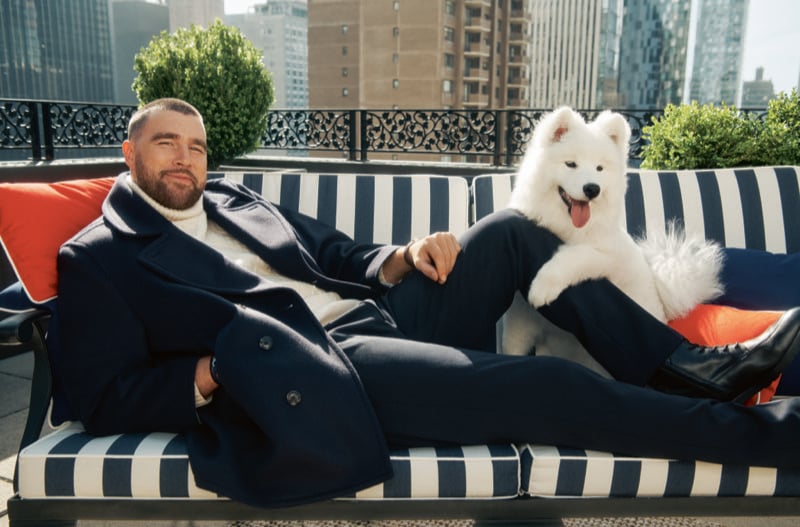 Travis Kelce reclines on a black-and-white striped terrace sofa next to a white Samoyed, wearing a double-breasted peacoat over a ribbed rollneck sweater, Manhattan towers behind him, for Tommy Hilfiger fall 2026