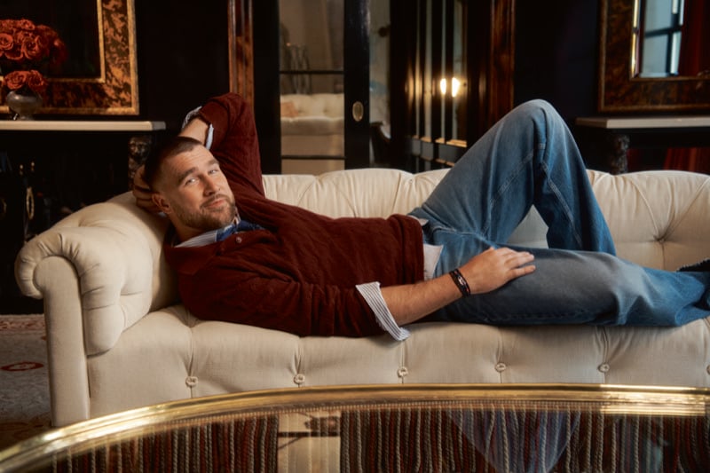Travis Kelce lies back on a pale tufted Chesterfield sofa in a dark Plaza sitting room with red roses, wearing a burgundy sweater over a striped shirt and light-wash jeans, for Tommy Hilfiger fall 2026