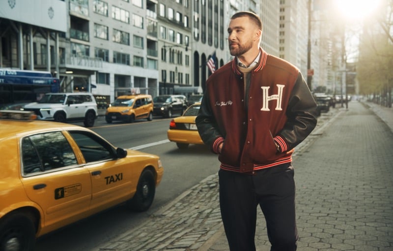 Travis Kelce crosses a Manhattan street past yellow taxis in a burgundy varsity jacket with leather sleeves and a large H patch, worn with dark trousers, for Tommy Hilfiger fall 2026
