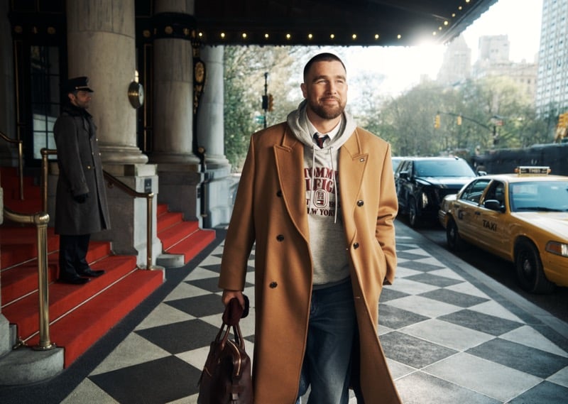 Travis Kelce walks beneath The Plaza's portico past a doorman on the red-carpeted steps, wearing a camel double-breasted overcoat over a gray Tommy Hilfiger hoodie, for fall 2026