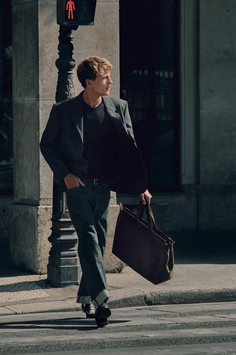 Vasko Luyckx crosses the street in a blazer and dark v-neck tee, tote bag in hand, for Zara's 90's Archive