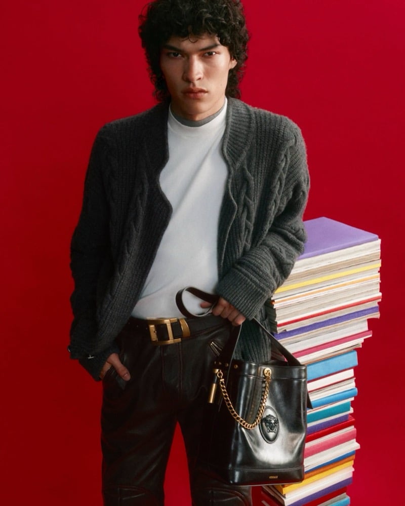 Jackson Roodman stands against a red backdrop in a gray Versace cable-knit cardigan and white top with dark leather trousers and a gold-buckle belt, holding a black leather Medusa chain bucket bag beside a tall stack of colorful books