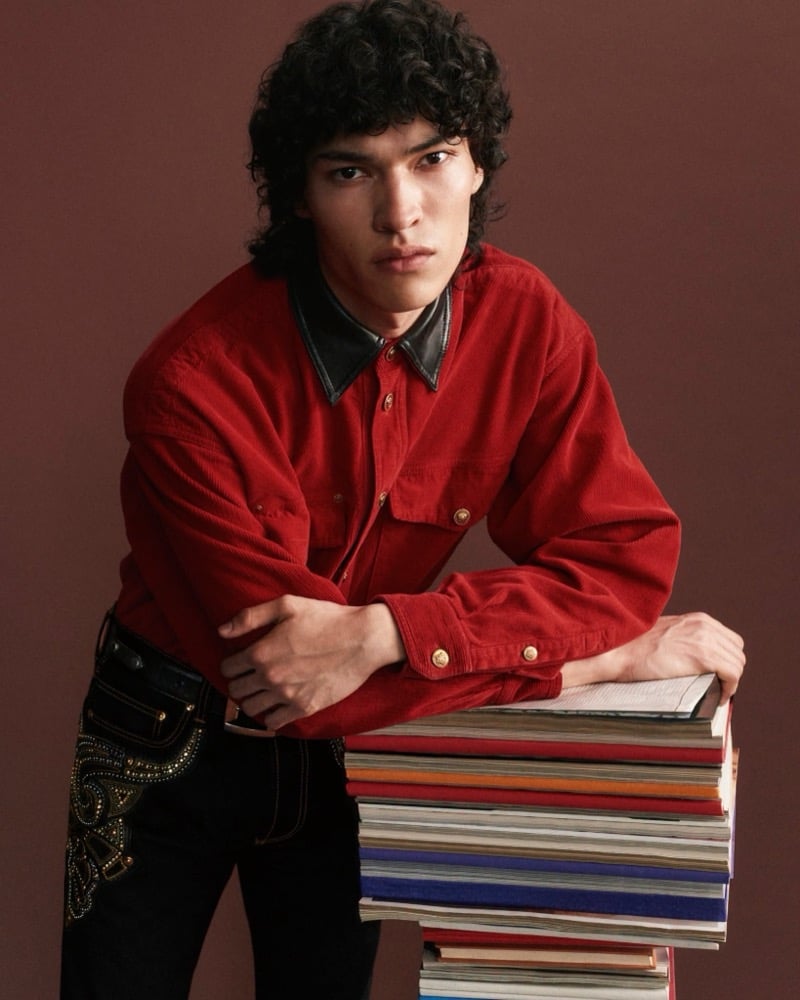 Jackson Roodman leans on a stack of books wearing a red Versace corduroy shirt with a leather collar and gold Medusa buttons, paired with black jeans embellished with gold studwork on the pocket