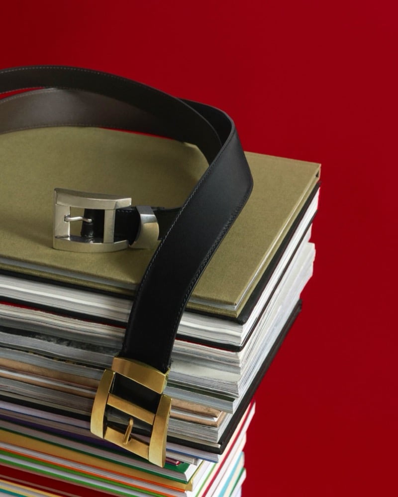 Two Versace leather belts, one with a brushed silver buckle and one with a gold buckle, draped over a stack of books against a red backdrop
