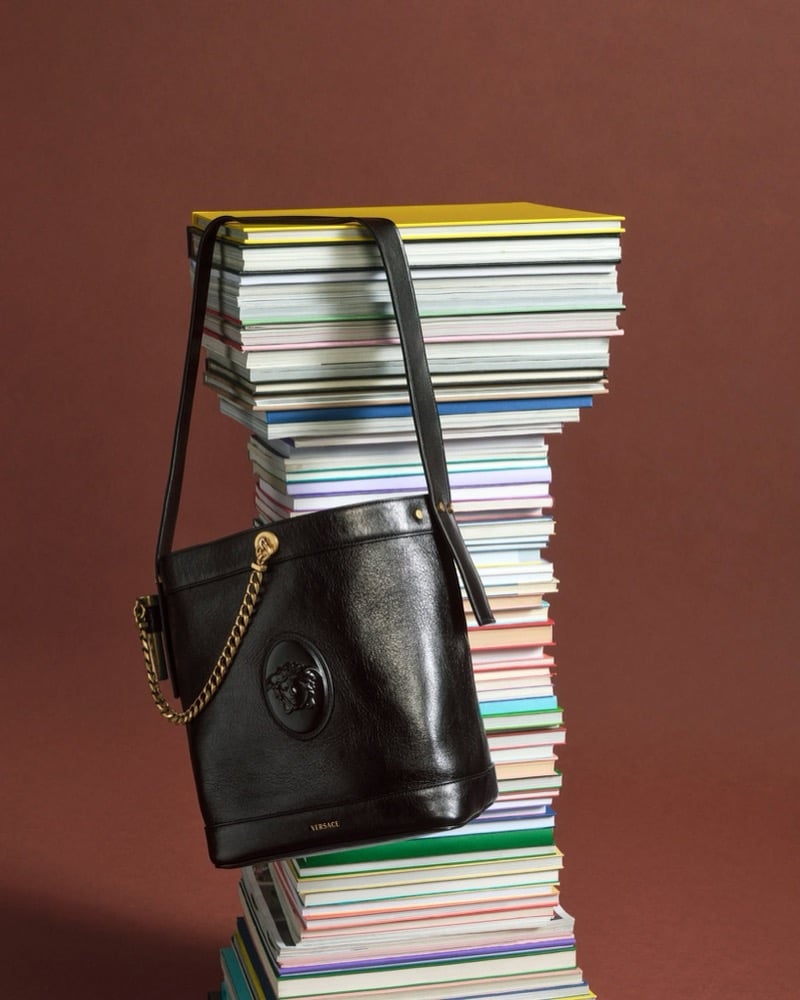 A black leather Versace Medusa bucket bag with a gold chain-and-leather strap, hanging from a tall stack of books against a brown backdrop