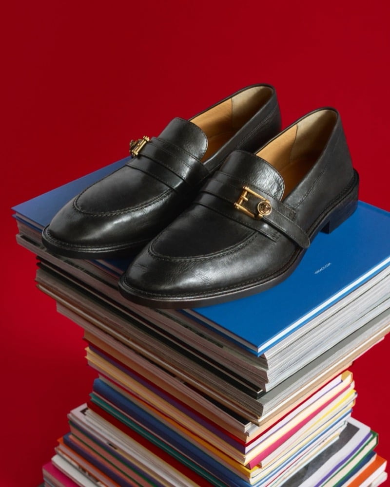 A pair of black Versace leather loafers with gold Medusa hardware on the strap, set on a stack of magazines against a red backdrop
