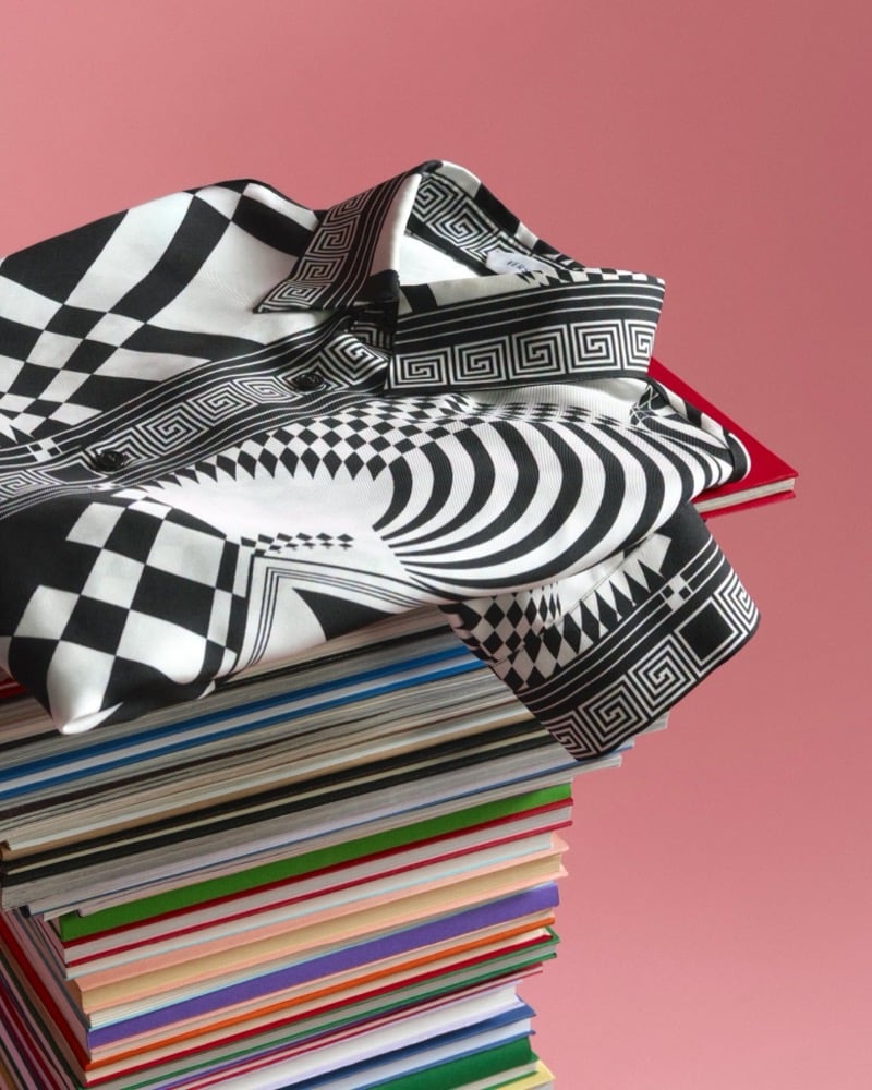 A folded Versace shirt in a black and white optical checkerboard print with Greek key trim, resting on a stack of books against a pink backdrop