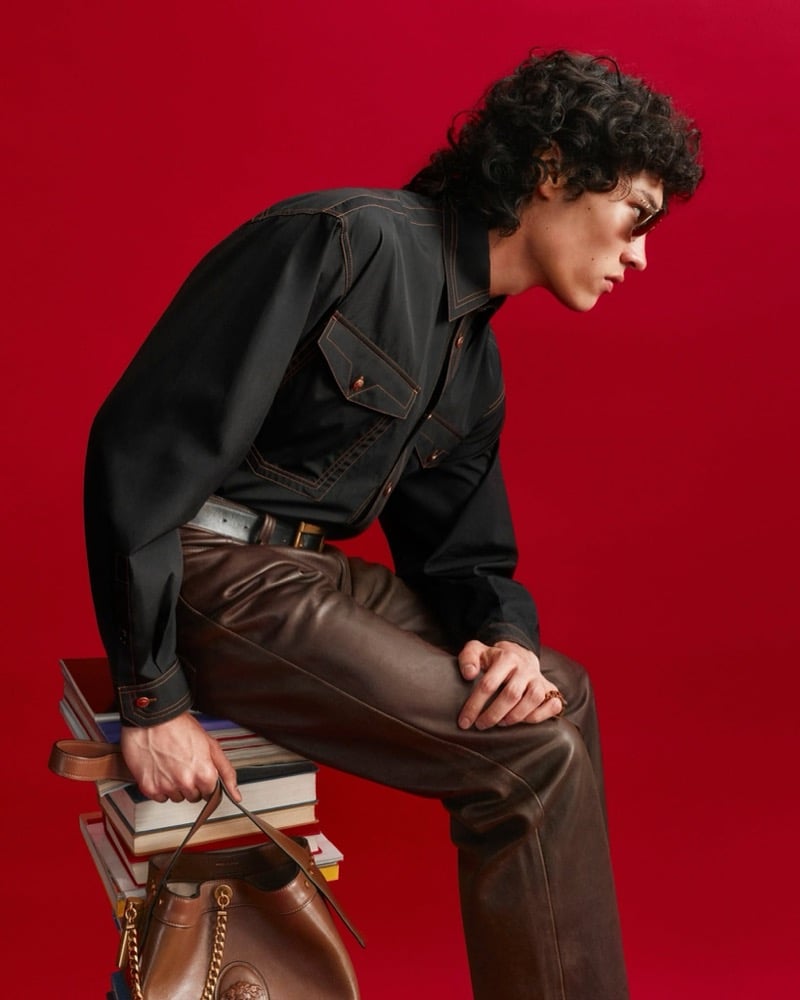 Jackson Roodman sits on a stack of books against a red backdrop in a black Versace overshirt with contrast topstitching and patch pockets, brown leather trousers, and sunglasses, a brown leather Medusa bucket bag at his feet
