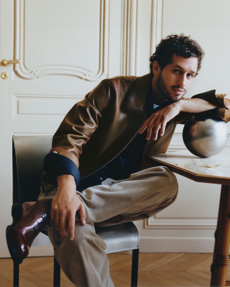 Victor Belmondo seated with his legs crossed in a brown leather jacket, taupe pleated trousers, and burgundy leather lace-up boots, leaning on a wood table with a metal sphere in a Parisian apartment