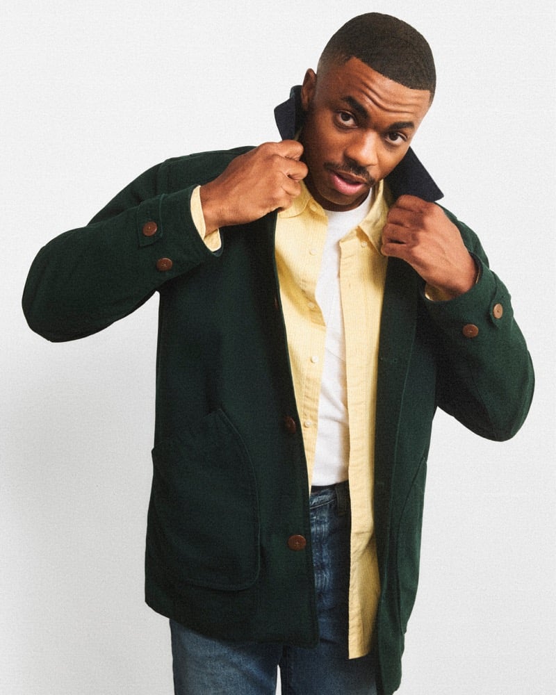Vince Staples pops the collar of a green J.Crew Barn Jacket worn over a yellow shirt and jeans