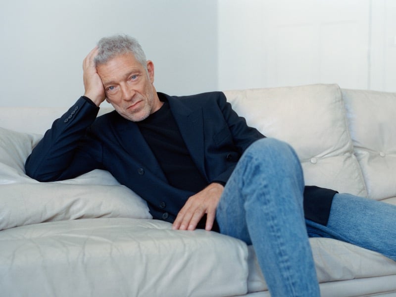 Vincent Cassel lounging on a leather sofa in a blazer, crewneck, and light-wash jeans for Mango