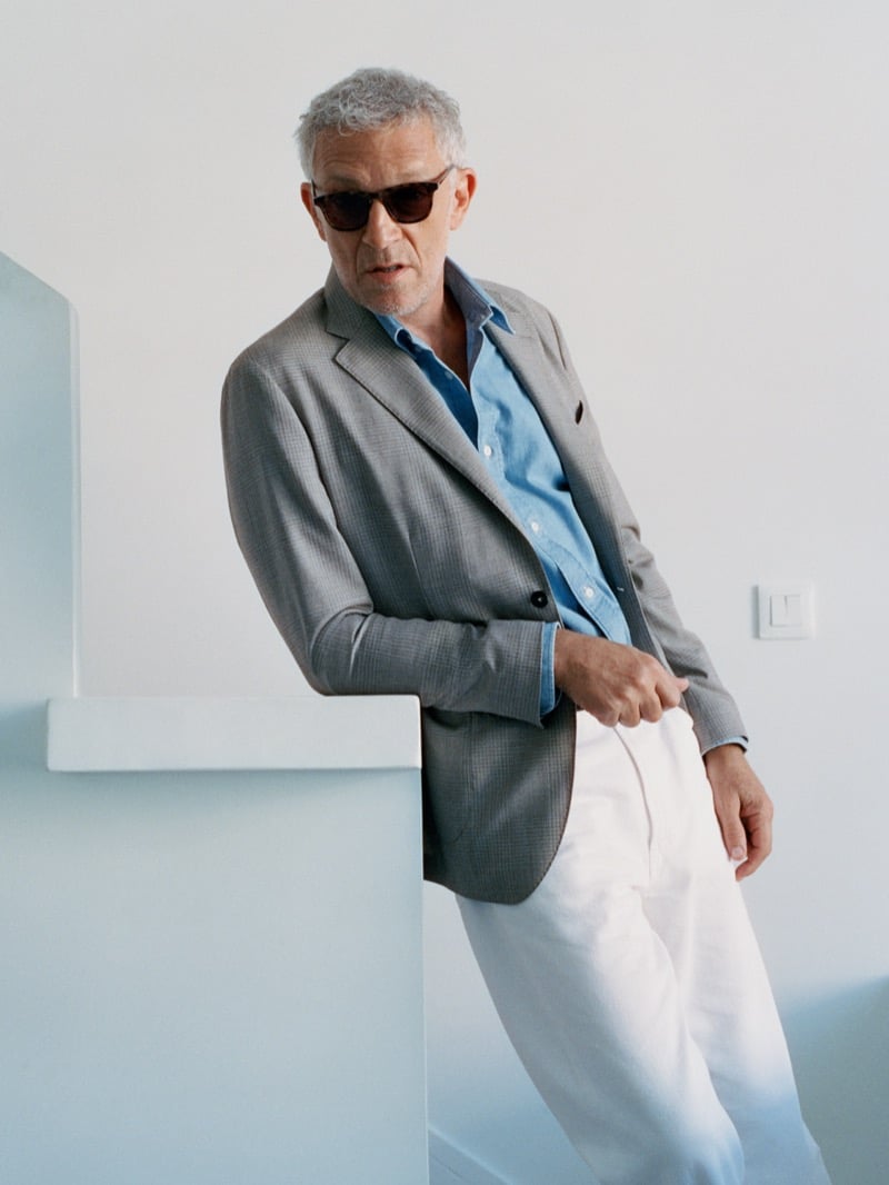Vincent Cassel in sunglasses, a gray check blazer, and a blue shirt, leaning on a stair wall for Mango