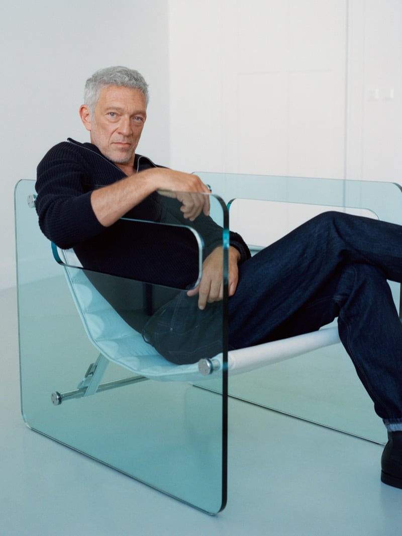 Vincent Cassel seated in a glass armchair wearing a collared knit sweater and dark-wash jeans for Mango