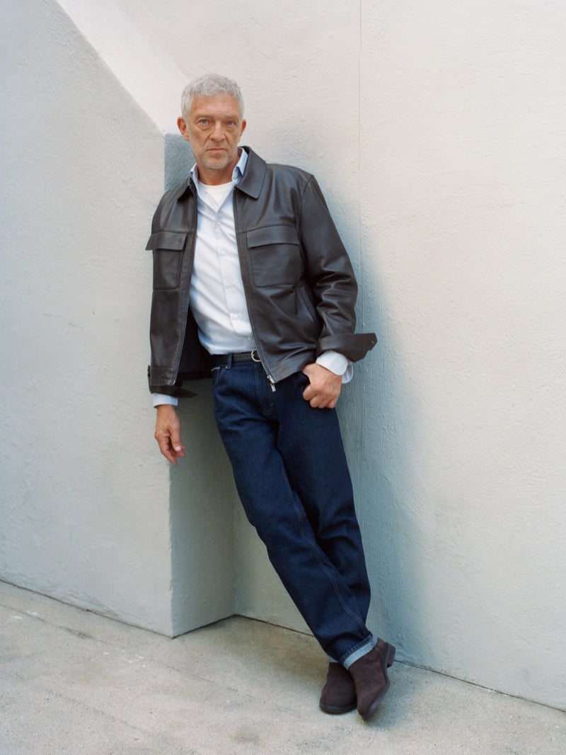 Vincent Cassel leans against a wall in a leather jacket, button-down shirt, dark-wash jeans, and suede boots for Mango