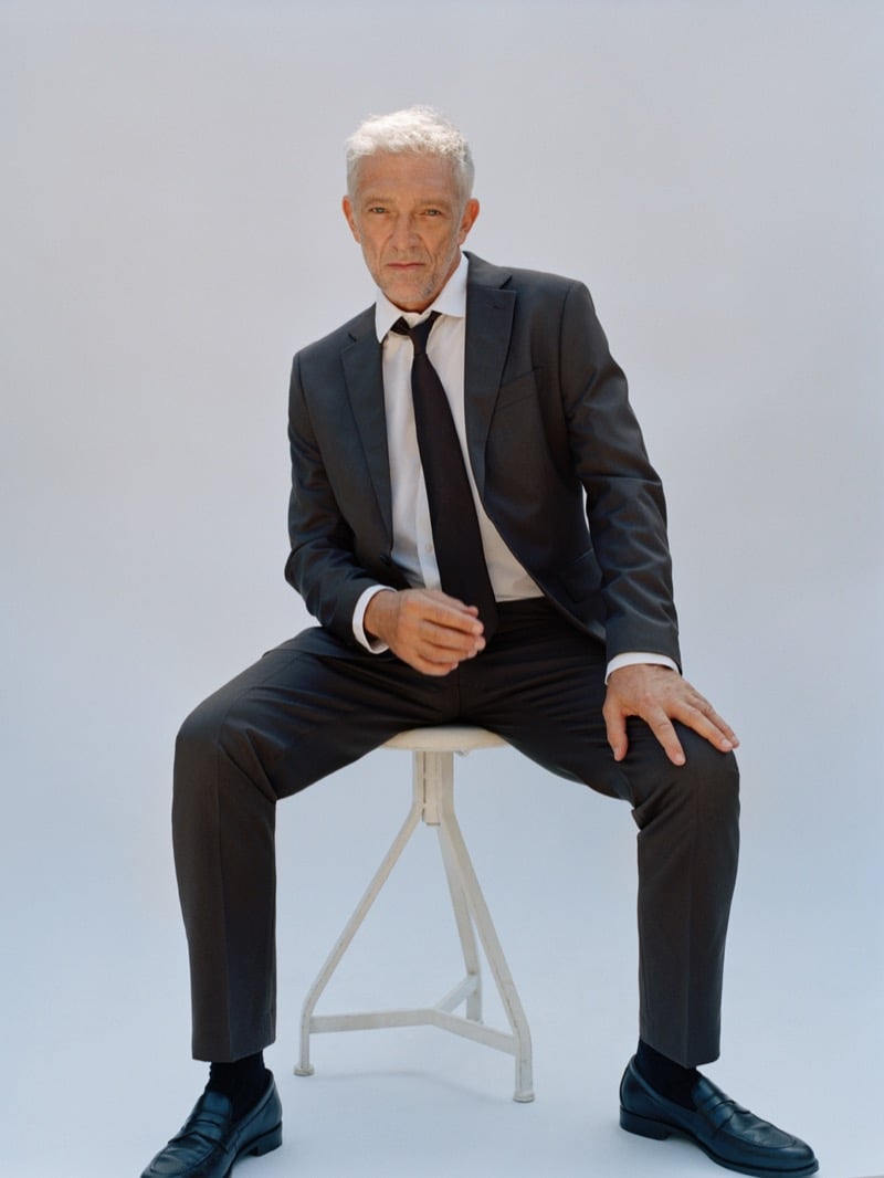 Vincent Cassel seated on a stool in a suit, dress shirt, tie, and penny loafers for Mango