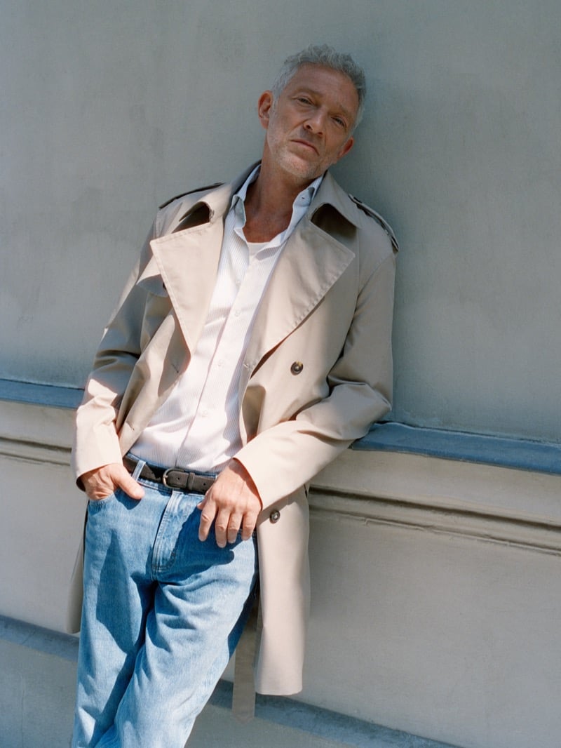 Vincent Cassel leans against a wall in a trench coat, open-collar striped shirt, and light-wash jeans for Mango