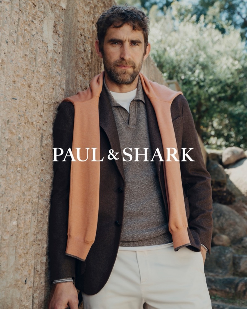 Will Chalker leaning against a textured stone wall in a brown blazer and gray knit polo with a salmon sweater draped over his shoulders