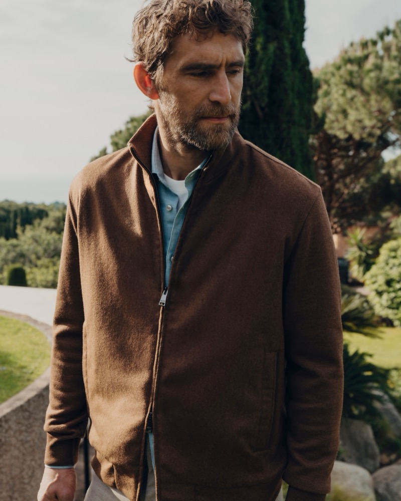 Will Chalker in a brown zip-front jacket over a light blue shirt with cypress trees and a garden behind him