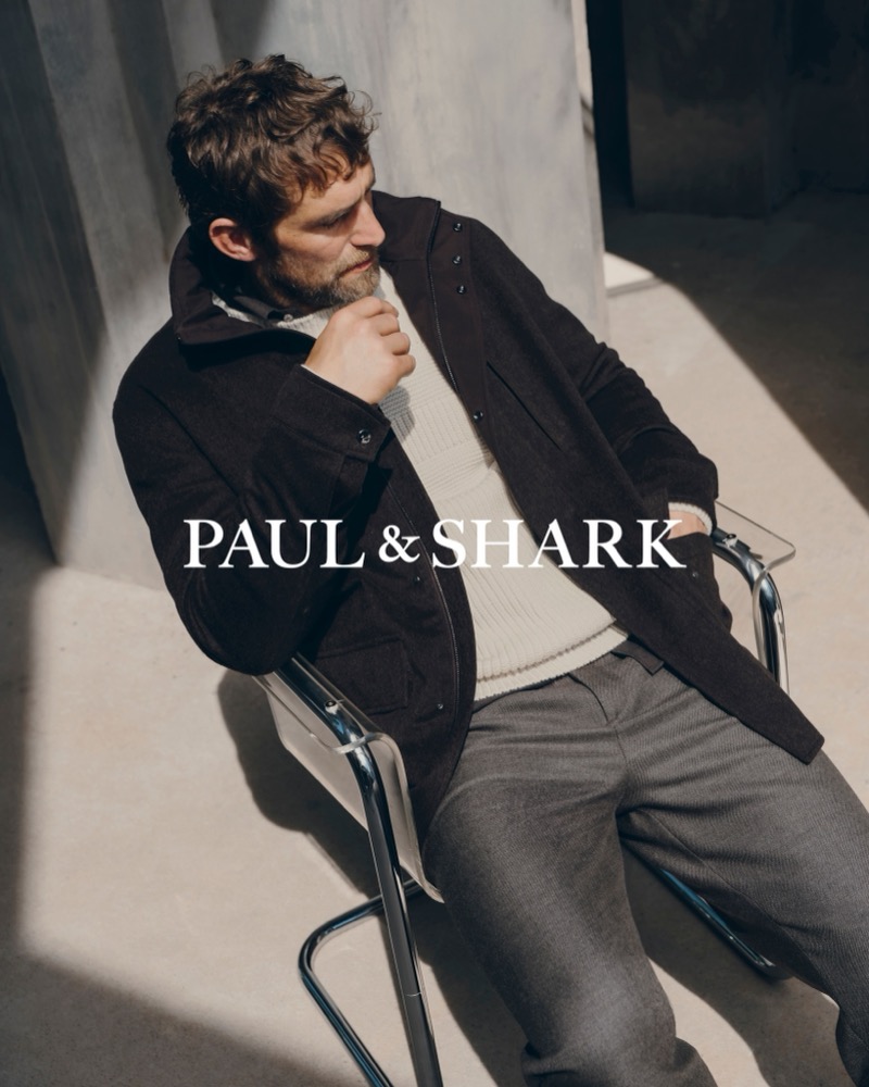 Will Chalker seated in a chrome-framed chair in a dark field jacket over a ribbed sweater and gray trousers, with sunlight across the concrete floor