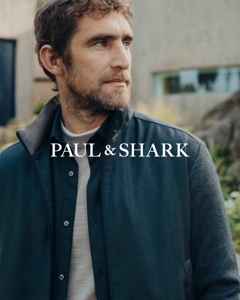 Will Chalker looking off to the side in a dark zip-front jacket with knit sleeves over a button-front shirt and gray crewneck