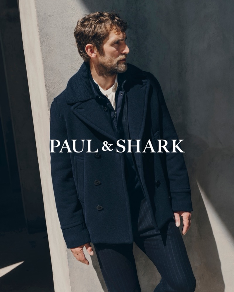 Will Chalker leaning against a sunlit concrete wall in a dark double-breasted peacoat and pinstripe trousers
