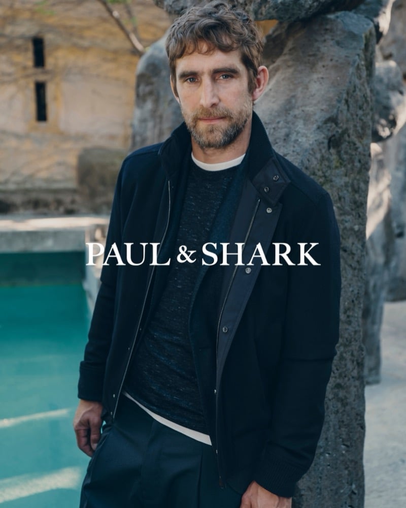 Will Chalker standing beside a stone column and turquoise pool in a dark zip jacket over a speckled sweater and pleated trousers