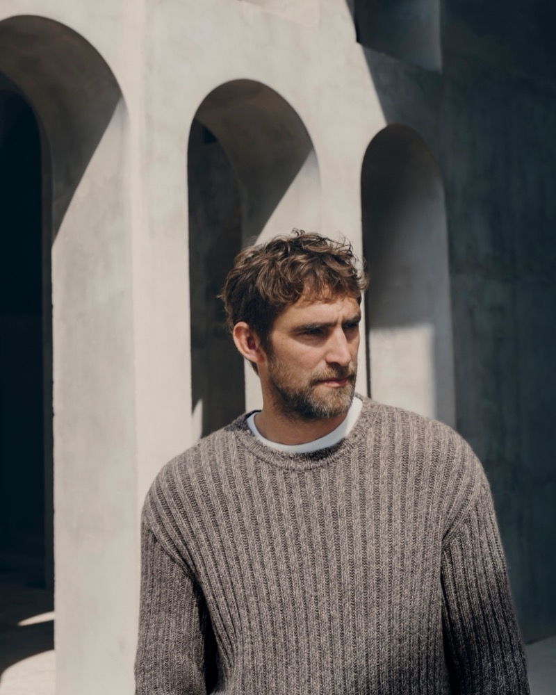 Will Chalker in a gray ribbed crewneck sweater standing in front of sunlit concrete arches