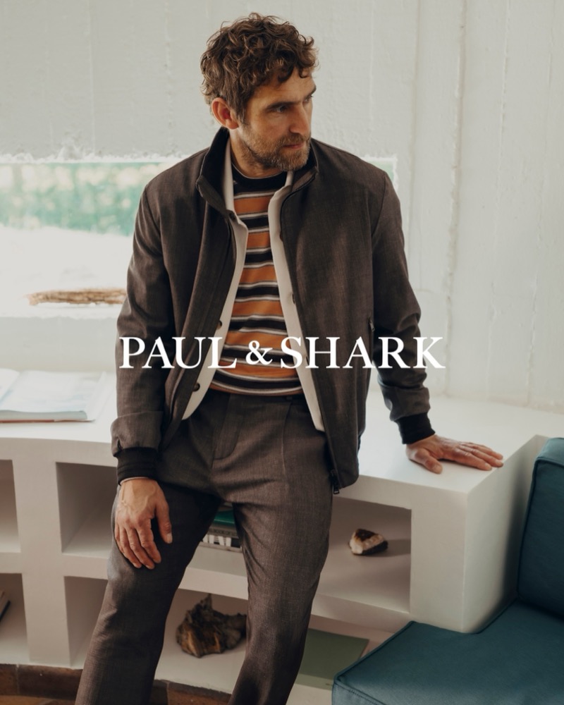 Will Chalker seated on a white shelf unit in a brown zip jacket over a striped knit and pleated brown trousers