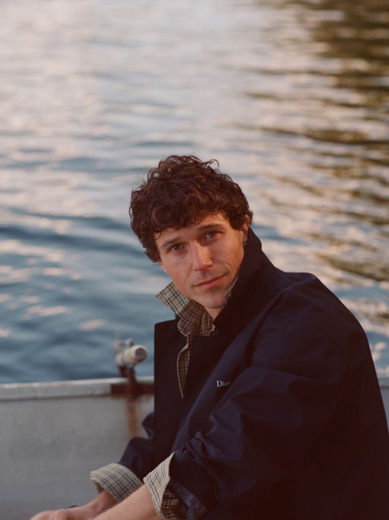 William Beck in a Dior logo jacket over a checked shirt sits in a rowboat with the lake behind